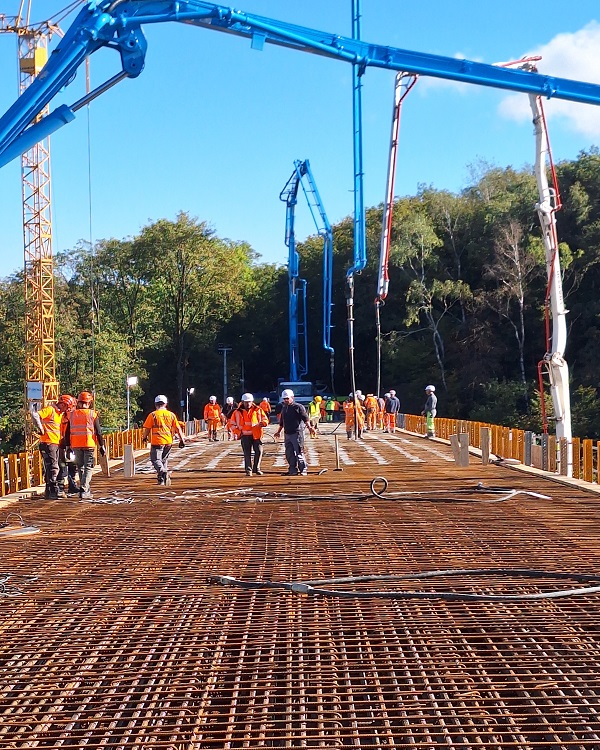 Das Bild zeigt eine Baustelle mit Bauarbeitern in orangefarbenen Westen auf einem Gitter aus Stahl. Im Hintergrund sind Baukräne und Bäume zu sehen.